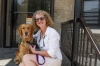 Lu Picard is sitting on a set of stairs smiling at the camera. Next to her is a golden retriever that is wearing a green assistance dog vest